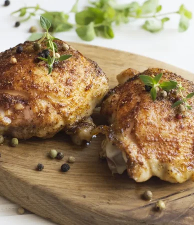 Two oven roasted chicken thighs with golden, crispy skin on a wooden board with fresh herbs
