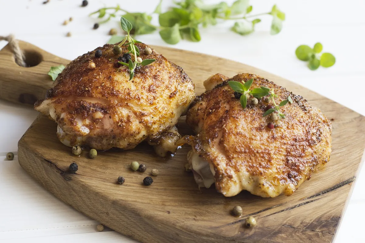 Two oven roasted chicken thighs with golden, crispy skin on a wooden board with fresh herbs