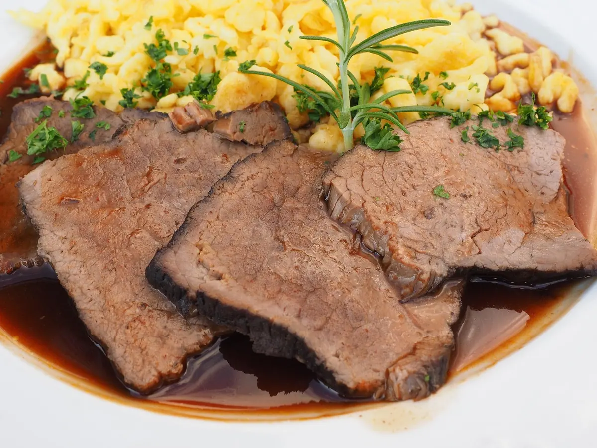 Slices of braised beef roast in a rich gravy with herbs and a side of spaetzle