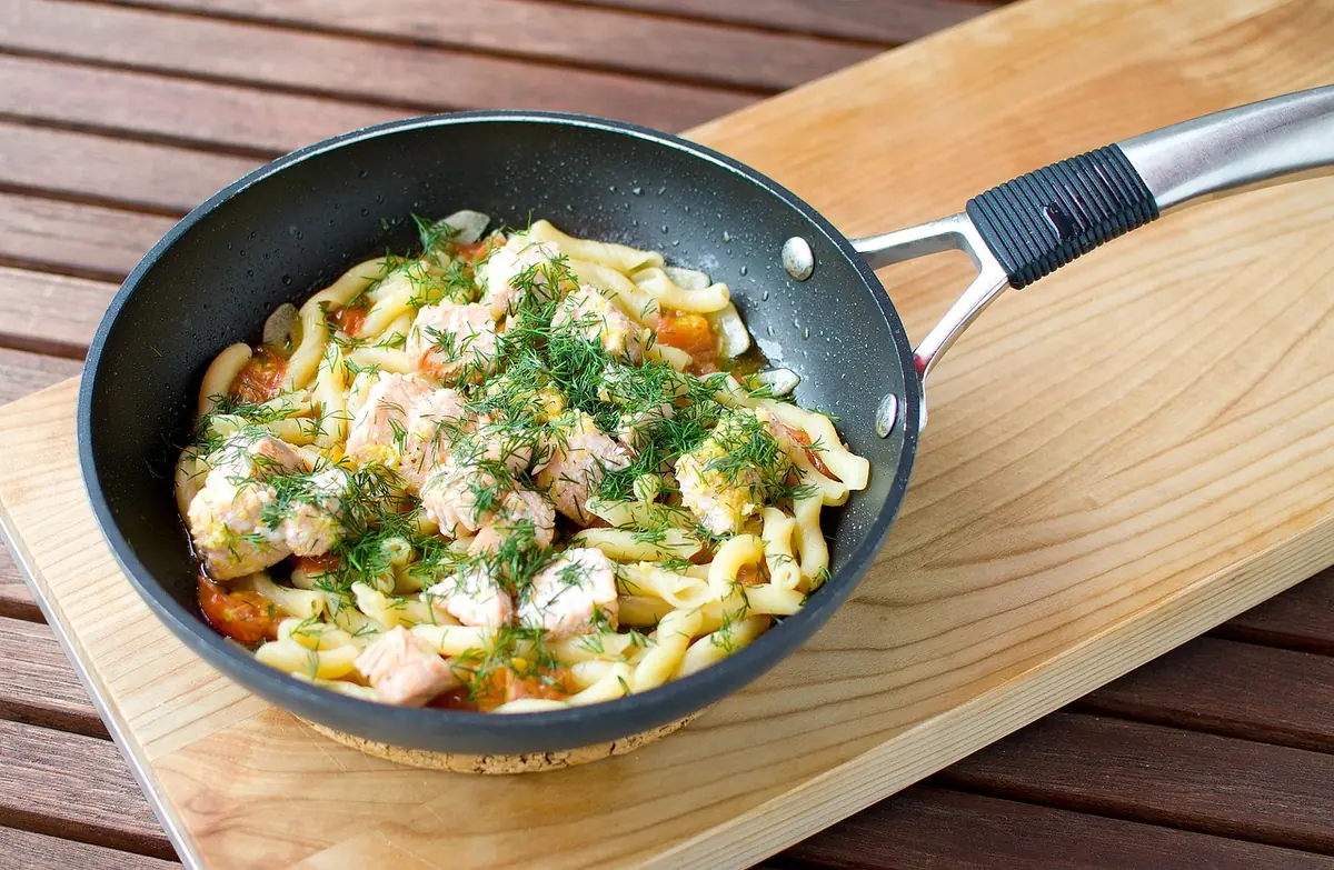 Skillet of pasta with flaked salmon and fresh dill on a wooden board
