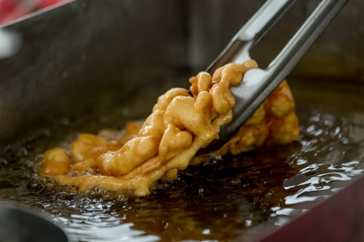 Funnel cake frying in hot oil, lifted with metal tongs