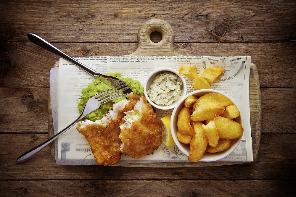 Crispy fried fish with a small bowl of tartar sauce and potato wedges on a wooden board