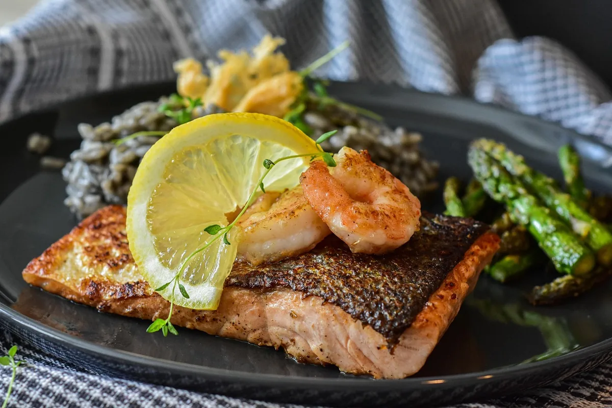 Crispy-skinned salmon fillet with a lemon slice, shrimp and asparagus on a dark plate
