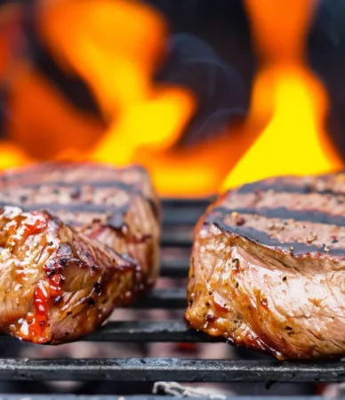 Two thick New York strip steak cuts searing on a grill grate with flames behind