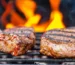 Two thick New York strip steak cuts searing on a grill grate with flames behind