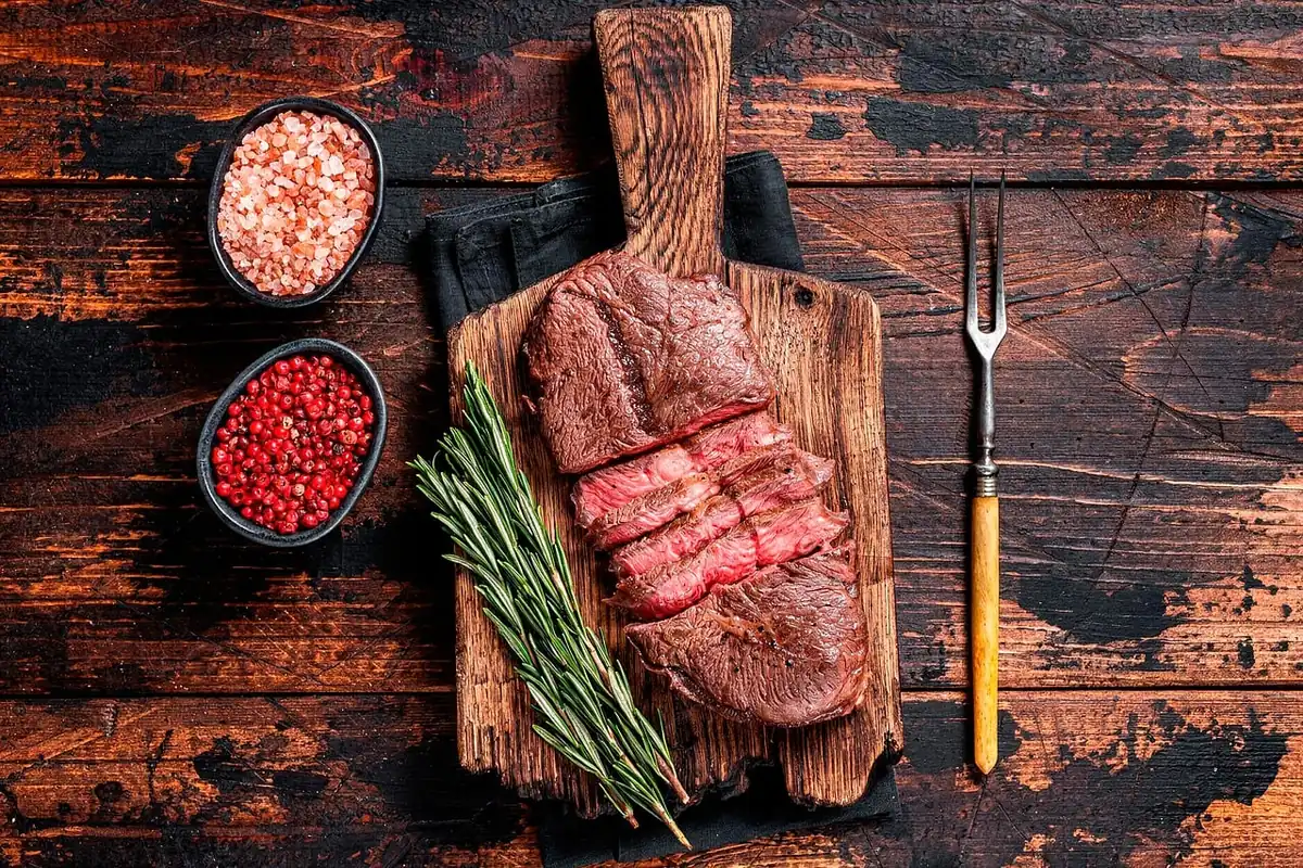 Sliced medium-rare steak on a wooden board with rosemary and peppercorns, ready for a steak sandwich