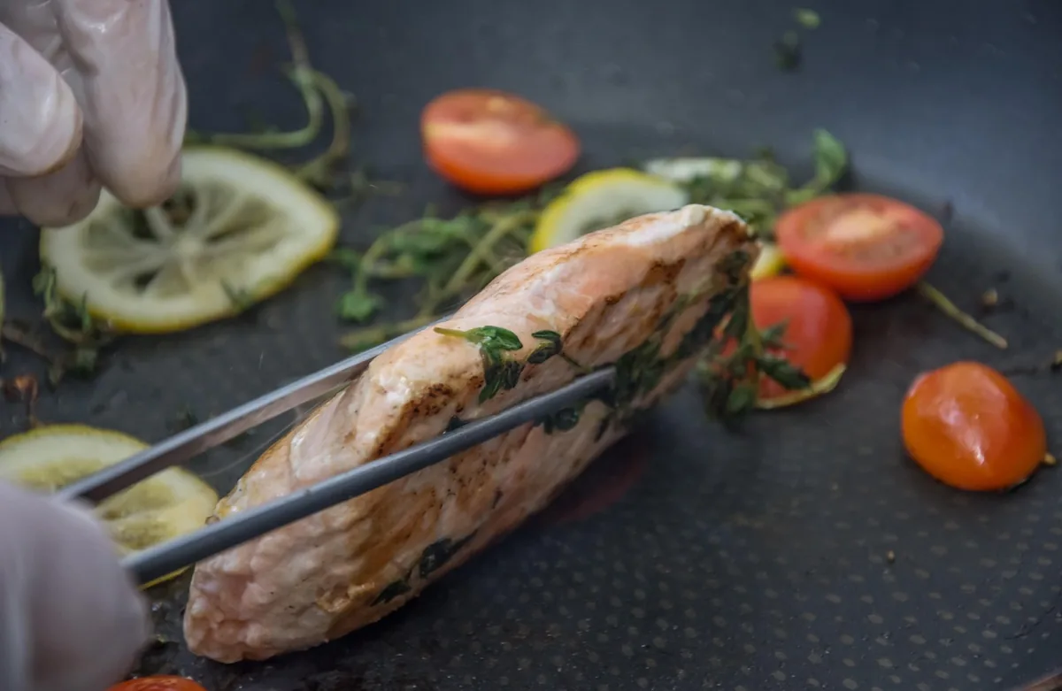 Salmon fillet being turned with tongs in a hot skillet with lemon slices and cherry tomatoes
