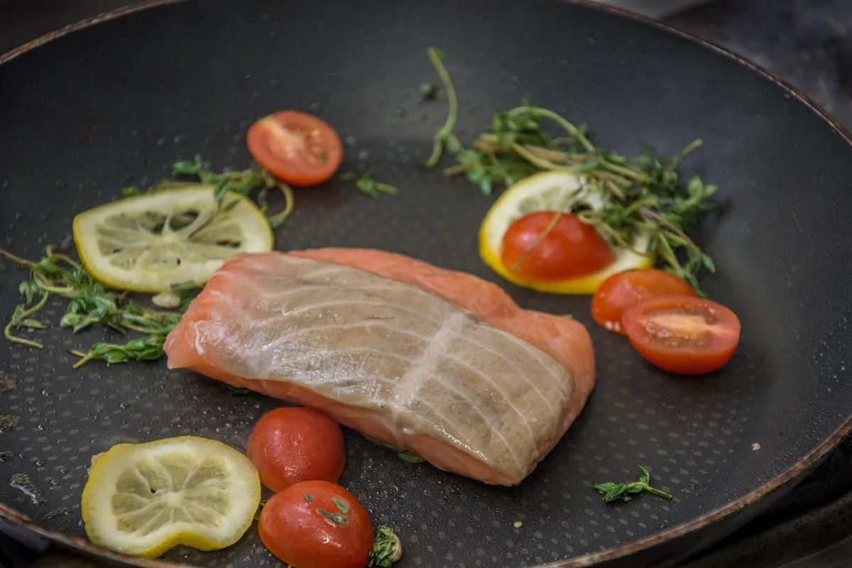 Salmon fillet cooking in a skillet with cherry tomatoes and lemon slices before glazing