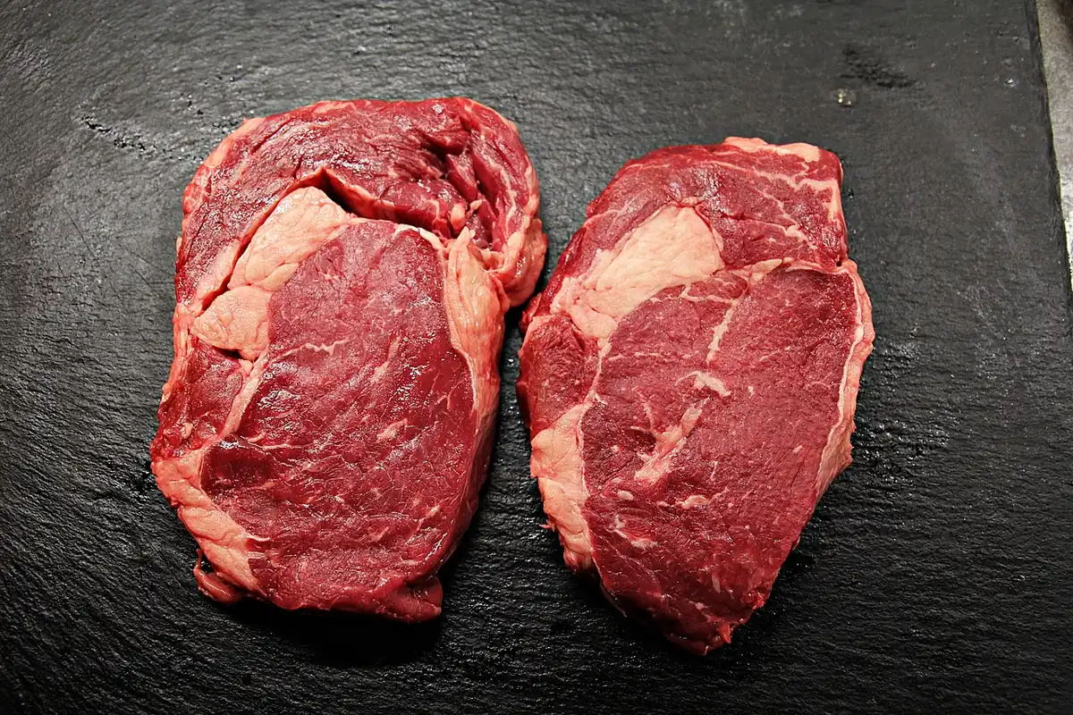 Two thick raw boneless ribeye steaks with fine marbling on a dark slate board