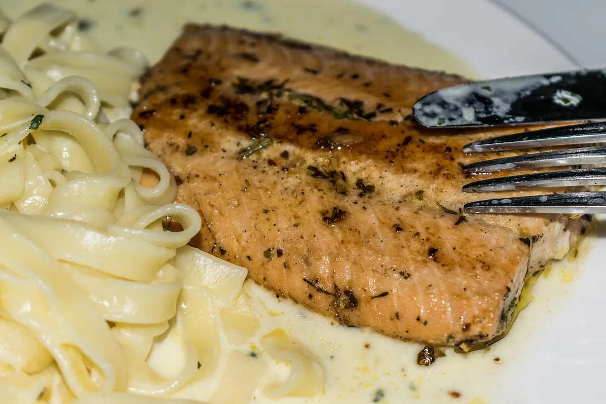 Pan-seared seasoned salmon fillet with herbs and black pepper next to creamy tagliatelle