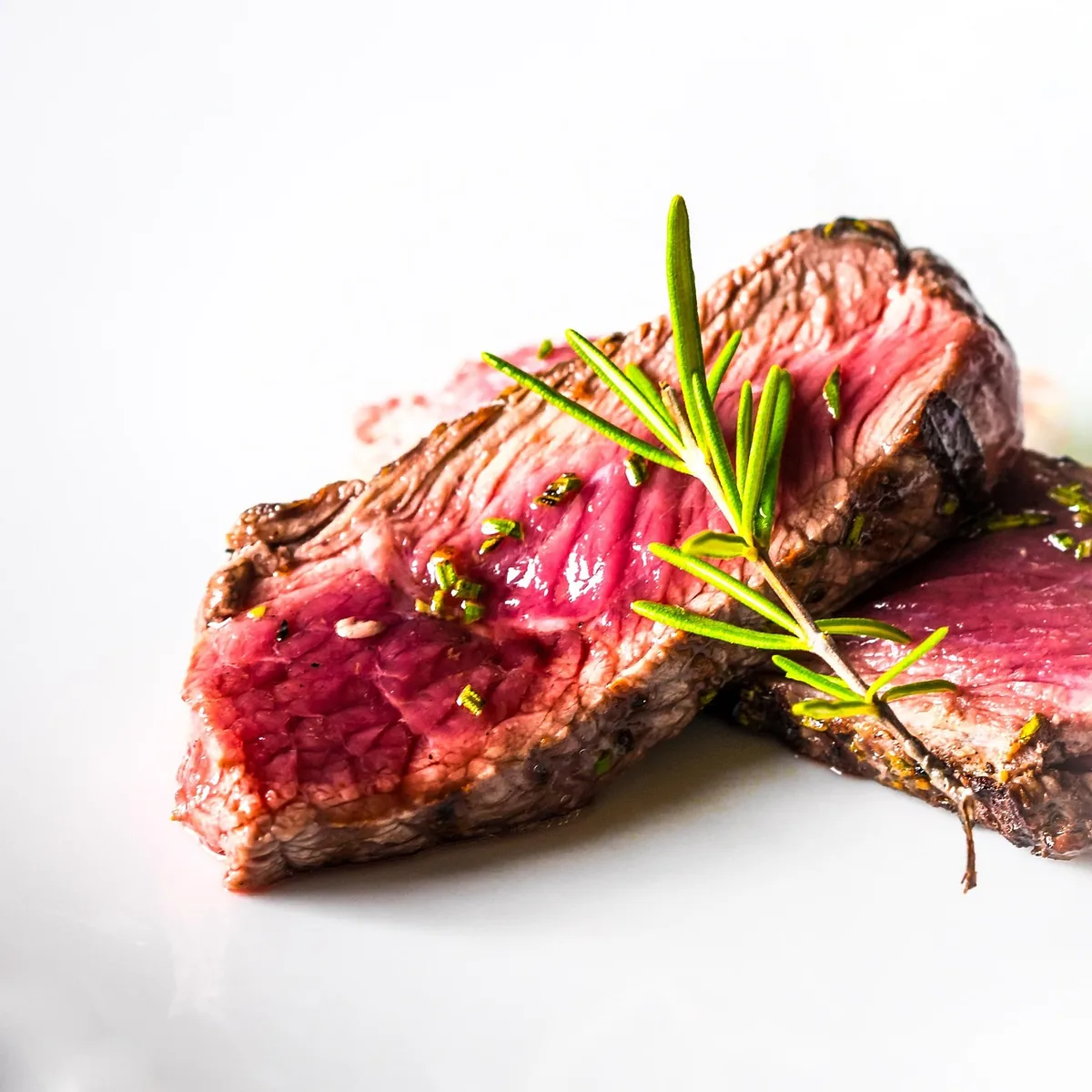 Sliced medium-rare grilled steak with a browned crust and a sprig of rosemary on a white plate