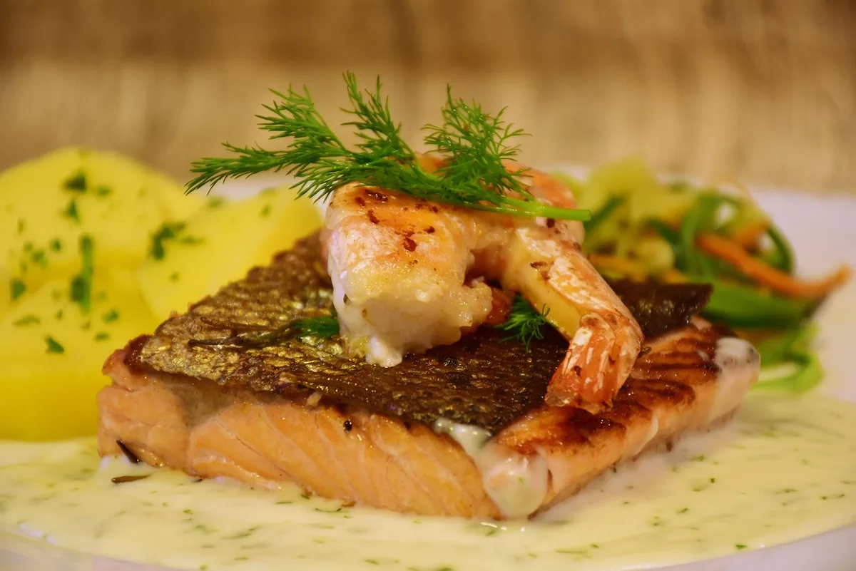 Seared salmon fillet topped with a grilled shrimp and fresh dill on a creamy dill sauce, with boiled potatoes