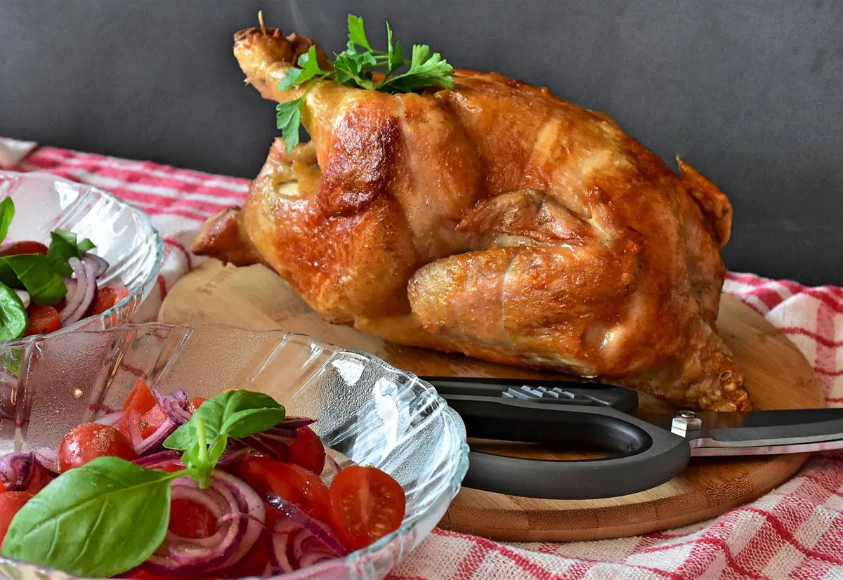 Roast whole chicken on a wooden board with poultry shears, ready to carve, next to a tomato salad