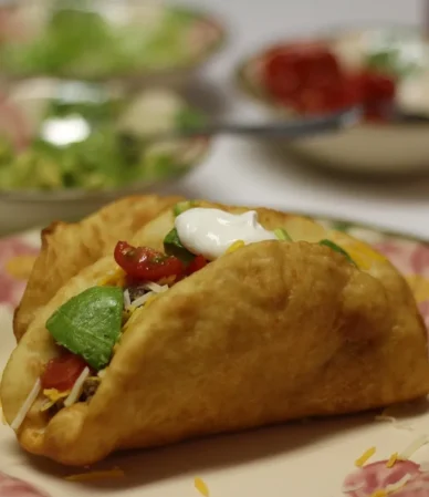 Crispy fried-shell taco filled with seasoned beef, tomato, avocado, cheese and sour cream on a floral plate, similar to ground beef tacos