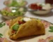 Crispy fried-shell taco filled with seasoned beef, tomato, avocado, cheese and sour cream on a floral plate, similar to ground beef tacos