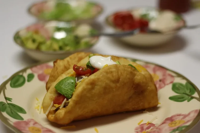 Crispy fried-shell taco filled with seasoned beef, tomato, avocado, cheese and sour cream on a floral plate, similar to ground beef tacos