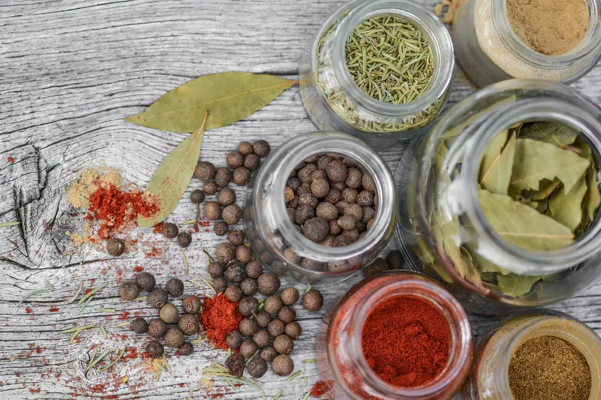 Glass jars of ground red chili spice, peppercorns, bay leaves and dried herbs on a wooden table for homemade taco seasoning
