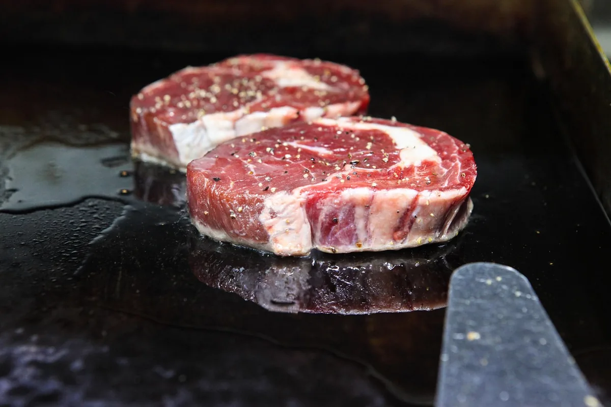 Two thick seasoned ribeye steaks searing on a hot black iron surface with a spatula, the final step of a reverse sear