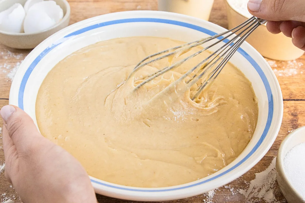 Hands whisking a smooth pale batter in a large bowl, the first step of a Dutch baby