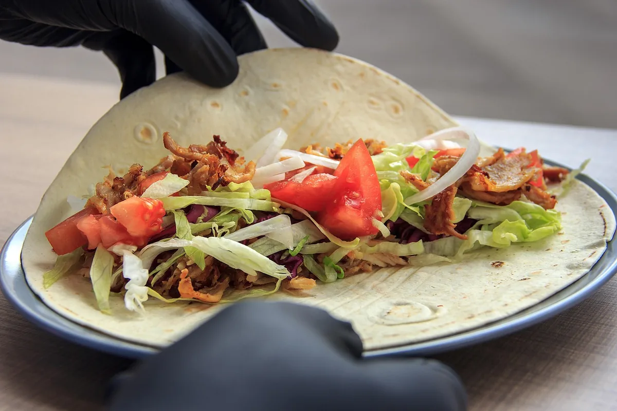 Gloved hands folding a flour tortilla over chicken, lettuce and tomato to roll a wrap