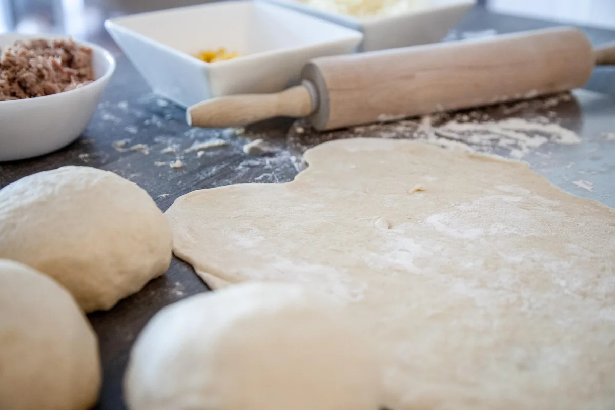 Pizza dough rolled out thin on a floured counter next to dough balls and a rolling pin, ready for chicken bakes