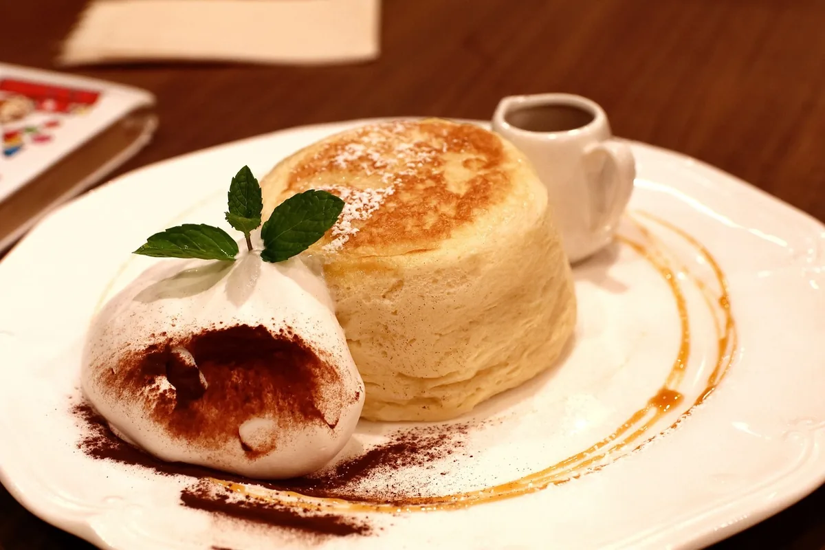 Tall golden souffle pancake dusted with sugar beside whipped cream and mint on a white plate, similar to Japanese souffle pancakes
