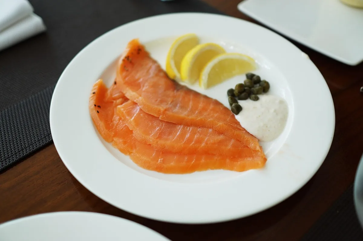 Sliced cold-smoked salmon on a white plate with lemon wedges, capers and a creamy dill sauce, ready to chop for dip