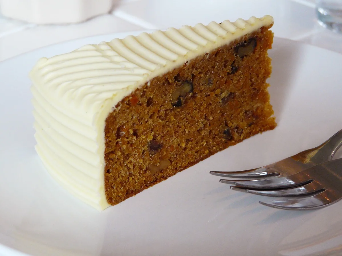 A slice of spice cake with nuts and thick cream cheese frosting on a white plate with a fork