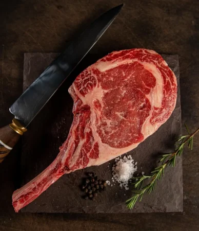 Raw marbled tomahawk steak with a long frenched rib bone on a dark board with a knife and rosemary