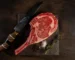 Raw marbled tomahawk steak with a long frenched rib bone on a dark board with a knife and rosemary