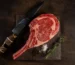 Raw marbled tomahawk steak with a long frenched rib bone on a dark board with a knife and rosemary