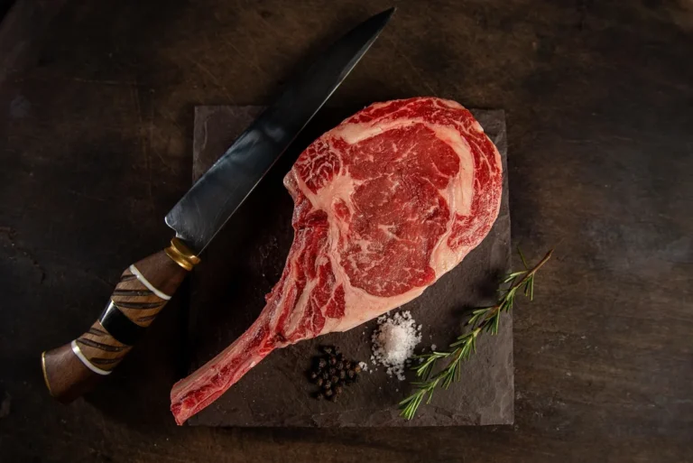 Raw marbled tomahawk steak with a long frenched rib bone on a dark board with a knife and rosemary