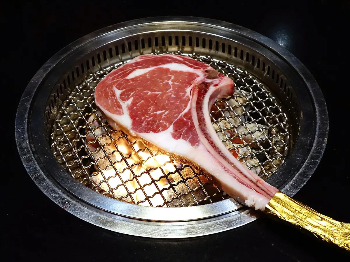 Thick tomahawk steak cooking on a round grill grate over glowing coals