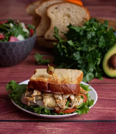 Chicken salad sandwich on toasted bread with lettuce, served with a bowl of salad, avocado and fresh herbs