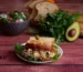 Chicken salad sandwich on toasted bread with lettuce, served with a bowl of salad, avocado and fresh herbs