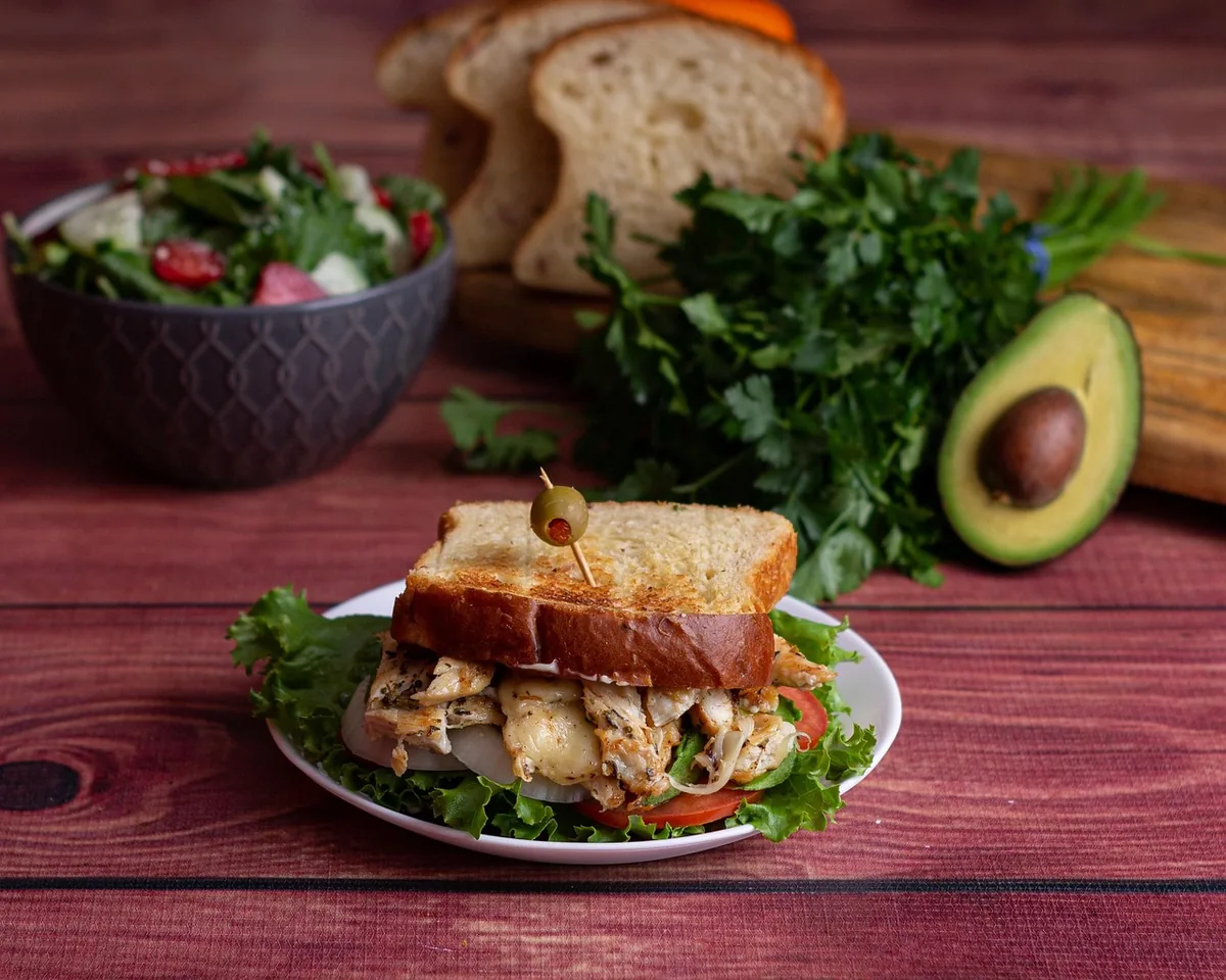 Chicken salad sandwich on toasted bread with lettuce, served with a bowl of salad, avocado and fresh herbs