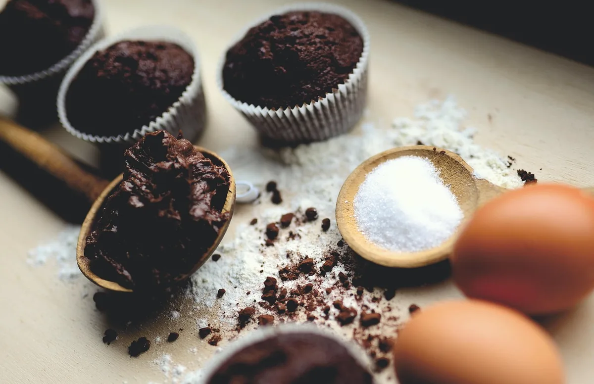 Baking ingredients on a counter: cocoa powder, sugar, flour and eggs, with chocolate muffins behind them