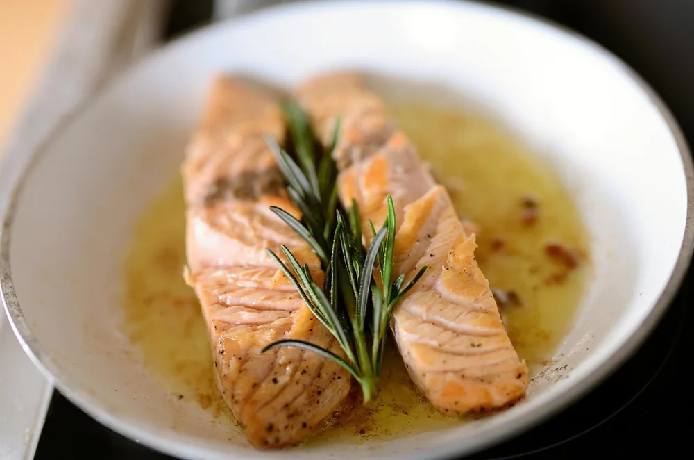 Two seared salmon pieces in a bowl of golden butter sauce with a sprig of rosemary