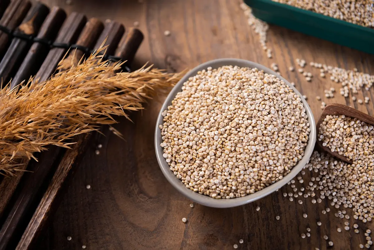 Bowl of uncooked white quinoa on a wooden table with a scoop of grains beside it