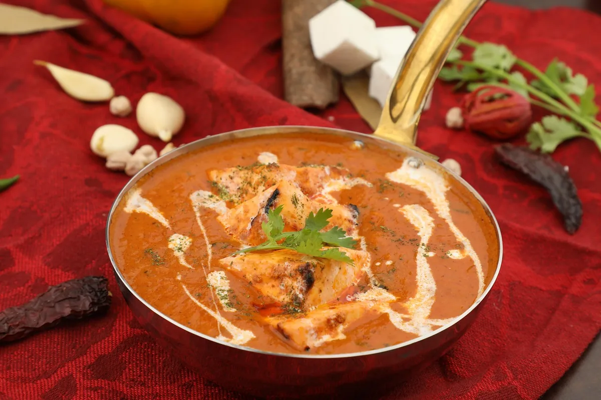 Paneer in a creamy orange tomato gravy in a copper pan, with raw paneer cubes behind