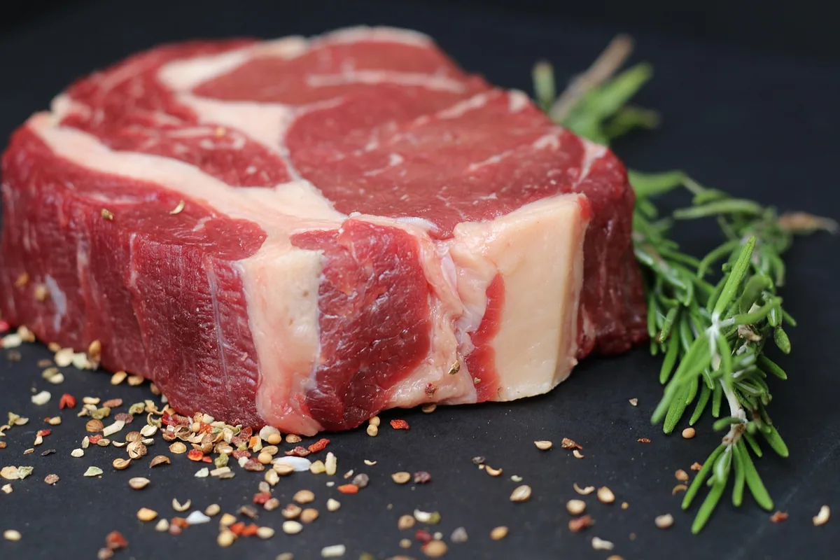 Thick raw ribeye steak with peppercorns, salt and rosemary on a dark board before seasoning